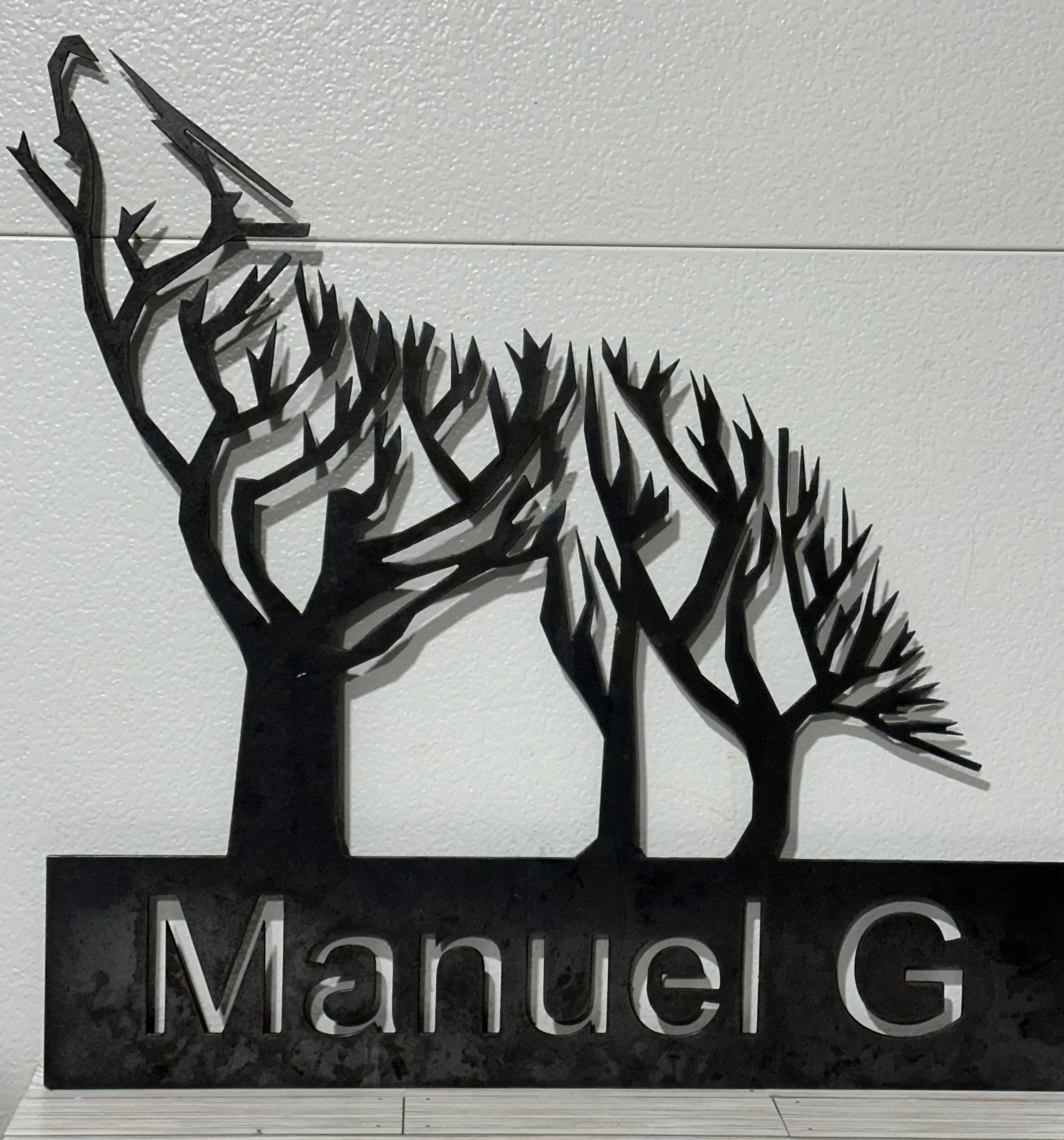 Metal wall art of wolf trees with branches, and the name 'Manuel G' beneath, mounted on a white wall.