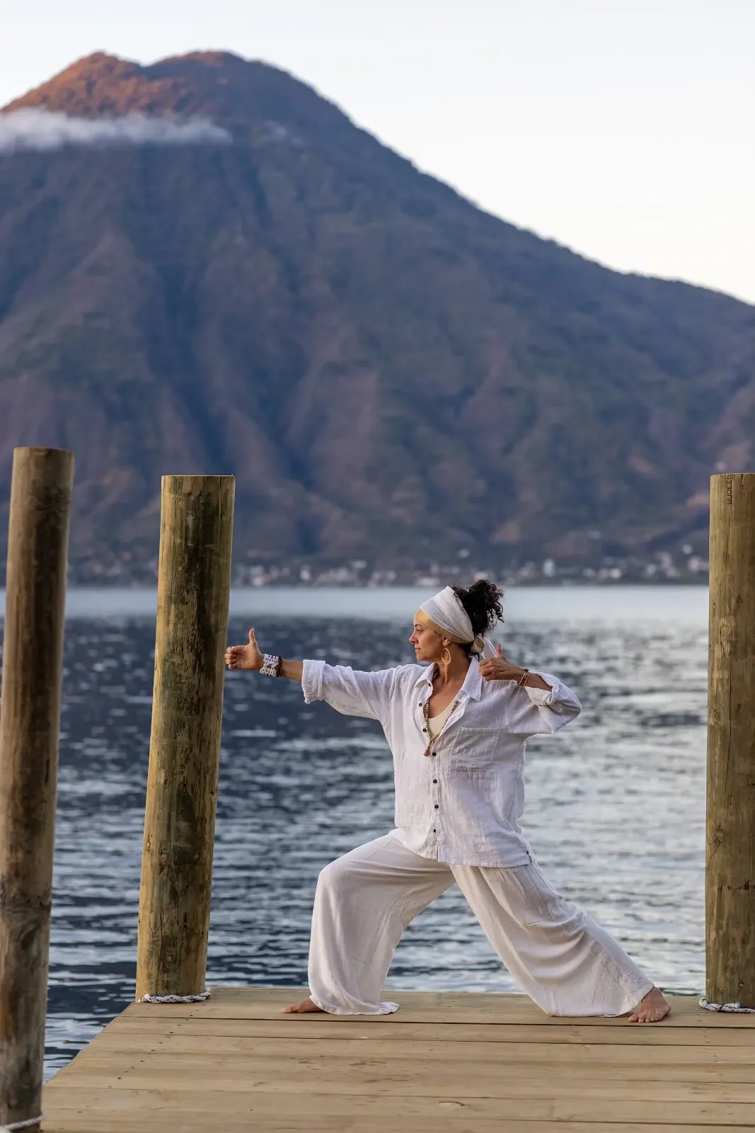 Woman sitting cross-legged in meditation pose, wearing white clothing and a white headwrap, with a large round mirror behind her, and meditation items around her.