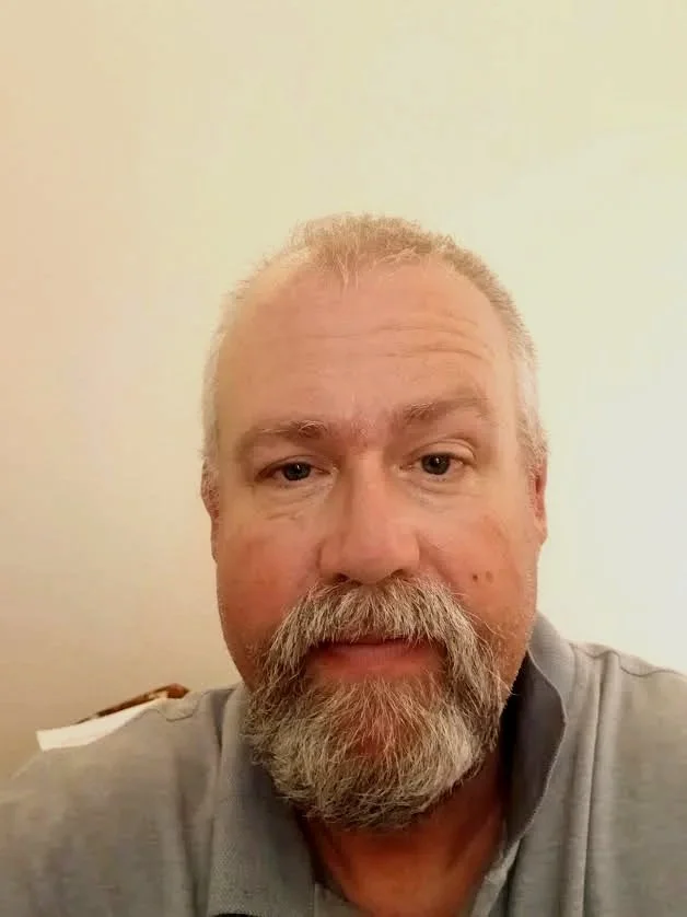 A woman with long, wavy strawberry blonde hair smiling while taking a selfie in a well-lit room with wooden floors and ceiling beams, and some photography equipment in the background.