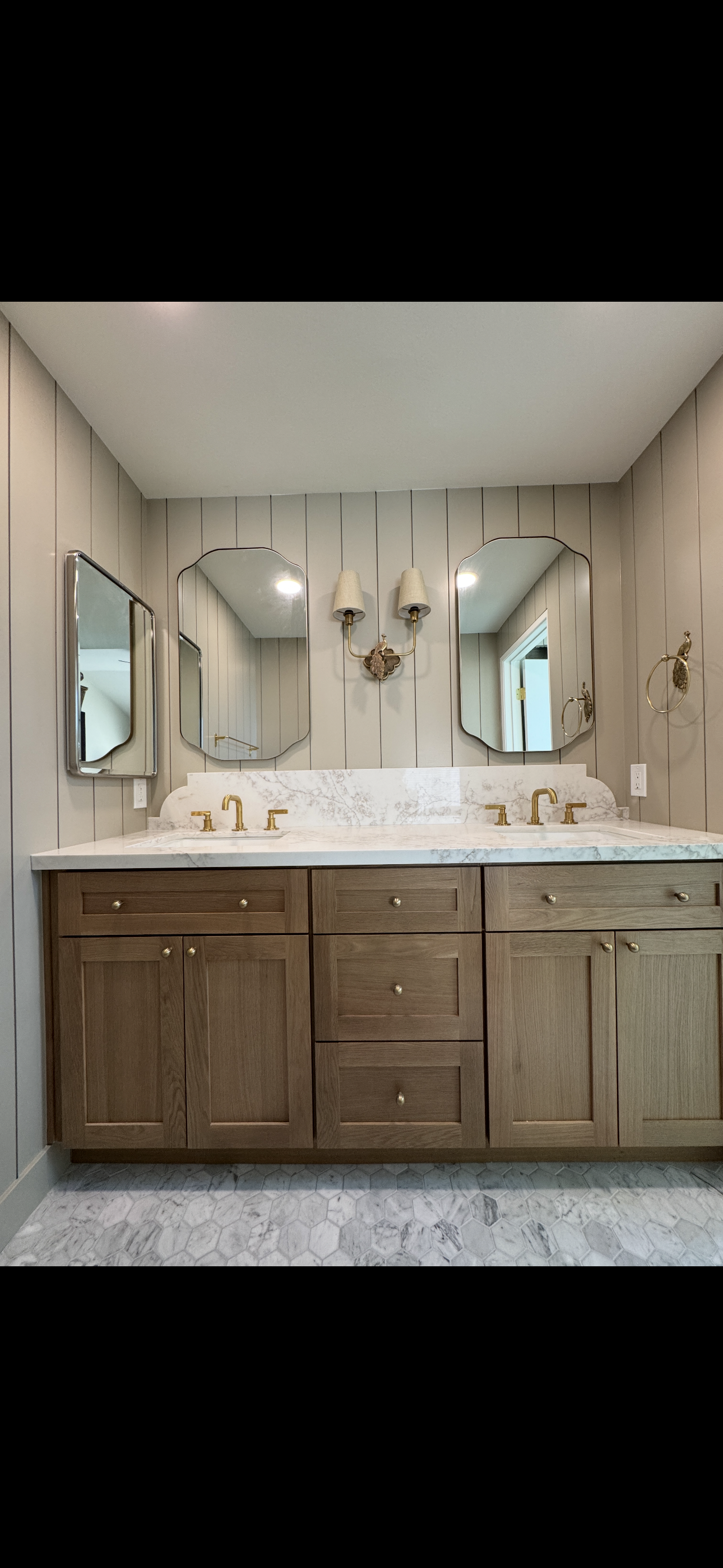 Bathroom vanity with a marble countertop, two sinks with gold fixtures, above three mirrors, beige panel walls, a double wall sconce, and hexagonal floor tiles.
