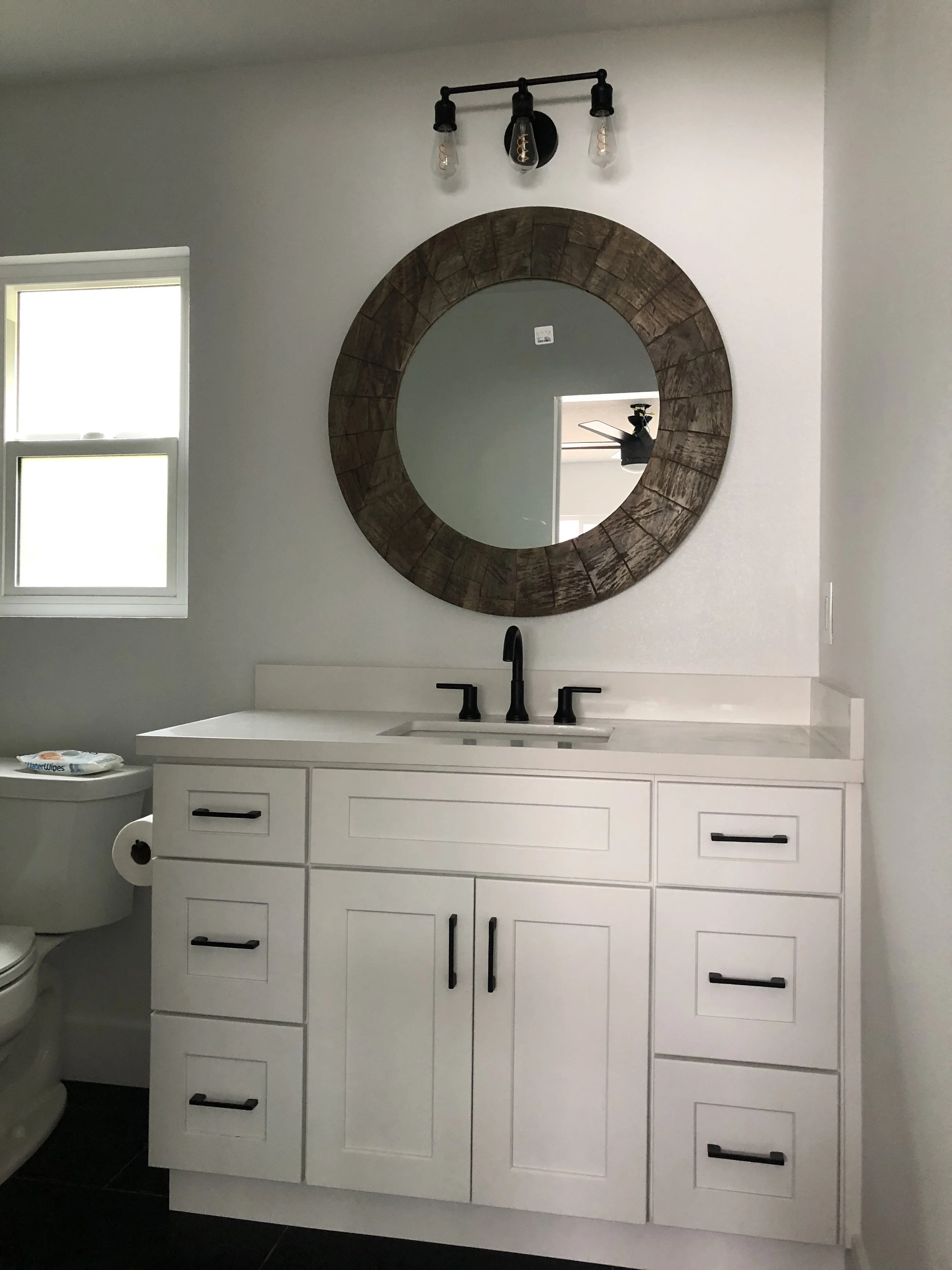 Bathroom vanity with a white cabinet, black handles, a black faucet, a round wooden-framed mirror, and a wall-mounted light fixture with three exposed bulbs.