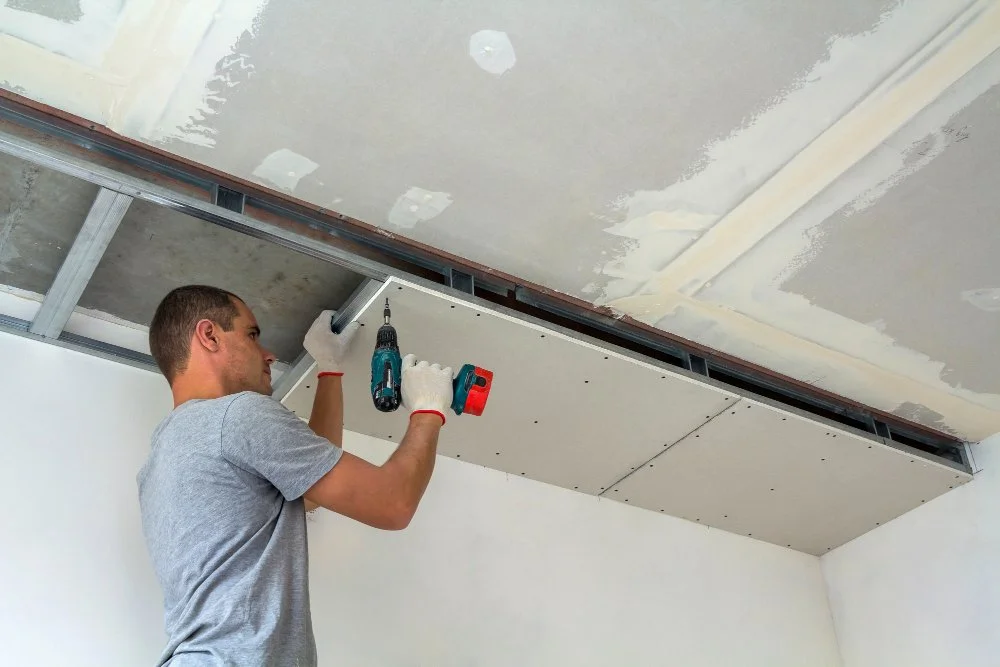 A man installing a suspended ceiling with drywall panels, using a power drill in a room with unfinished walls and ceiling.