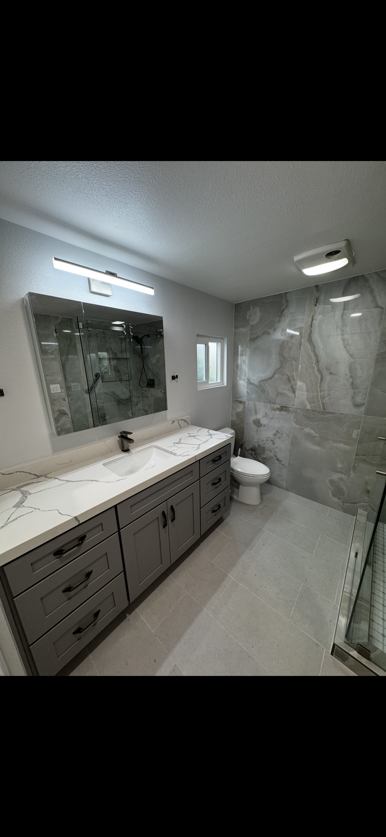 Modern bathroom remodel featuring a long gray vanity with marble countertop, large frameless mirror, recessed lighting, and a glass-enclosed shower with stone tile walls by Sam’s Renovations & Home Improvements.