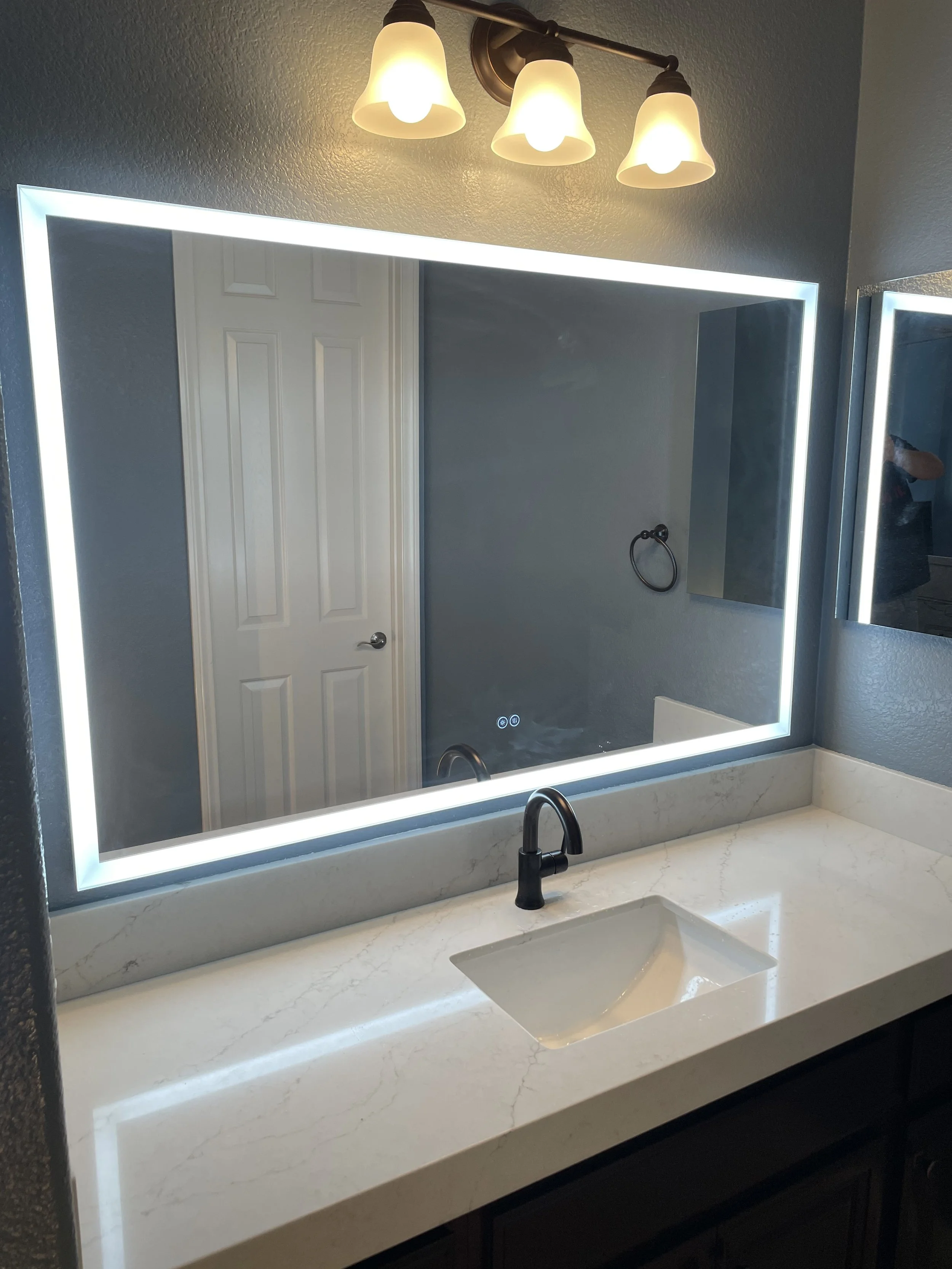 Modern bathroom vanity with quartz countertop, LED-lit mirror, black faucet, and overhead light fixture by Sam’s Renovations & Home Improvements.
