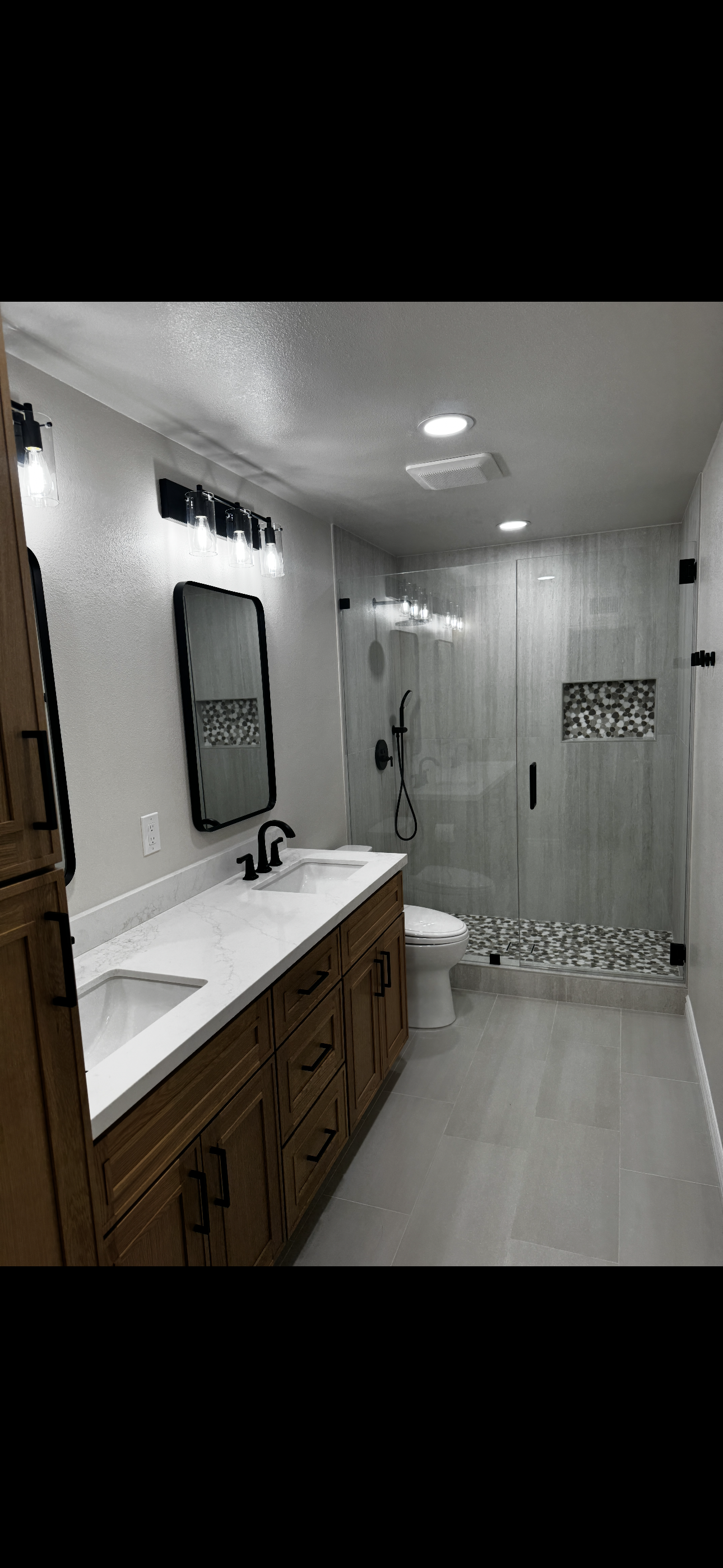 Modern bathroom remodel featuring a wood vanity with white countertop, black fixtures, large glass-enclosed shower with decorative tile accents, and recessed lighting by Sam’s Renovations & Home Improvements.