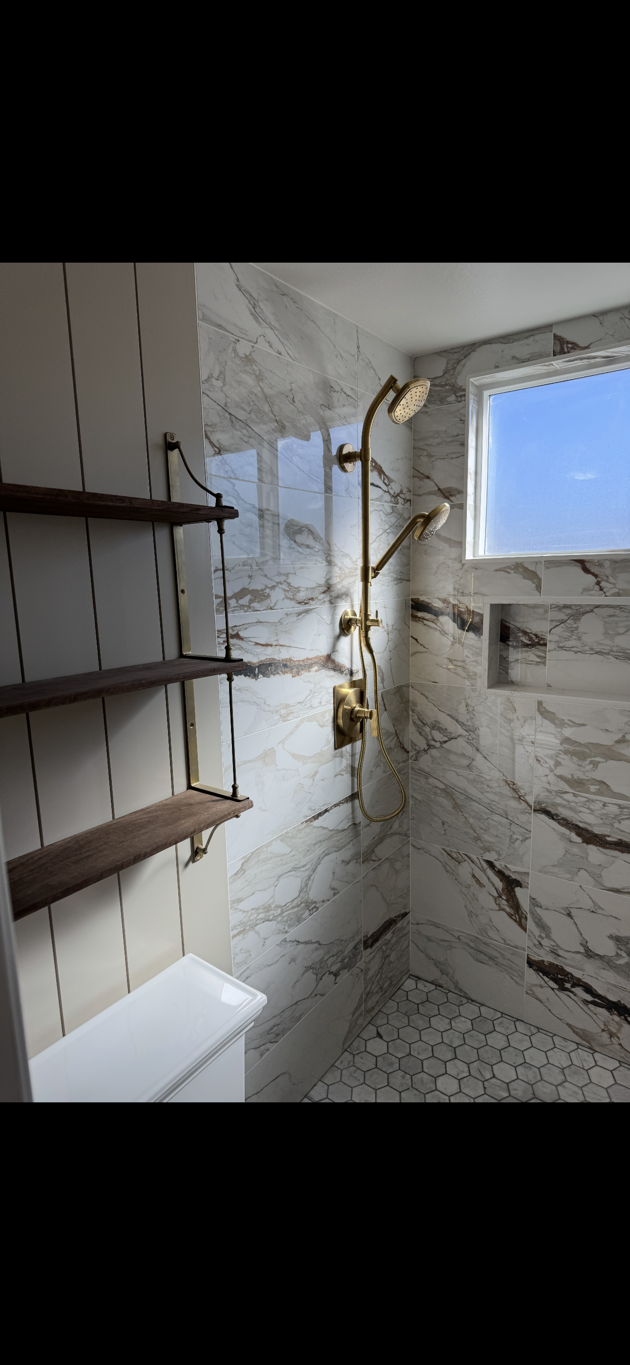 A modern walk-in shower with marble tile walls, a gold dual showerhead system, a small built-in shelf, a window with blue sky view, and wooden shelves on the left.