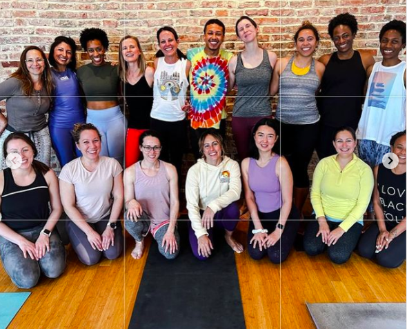 Group of 15 diverse women and one man posing together, some kneeling and some standing, in front of a brick wall with yoga mats on the floor.