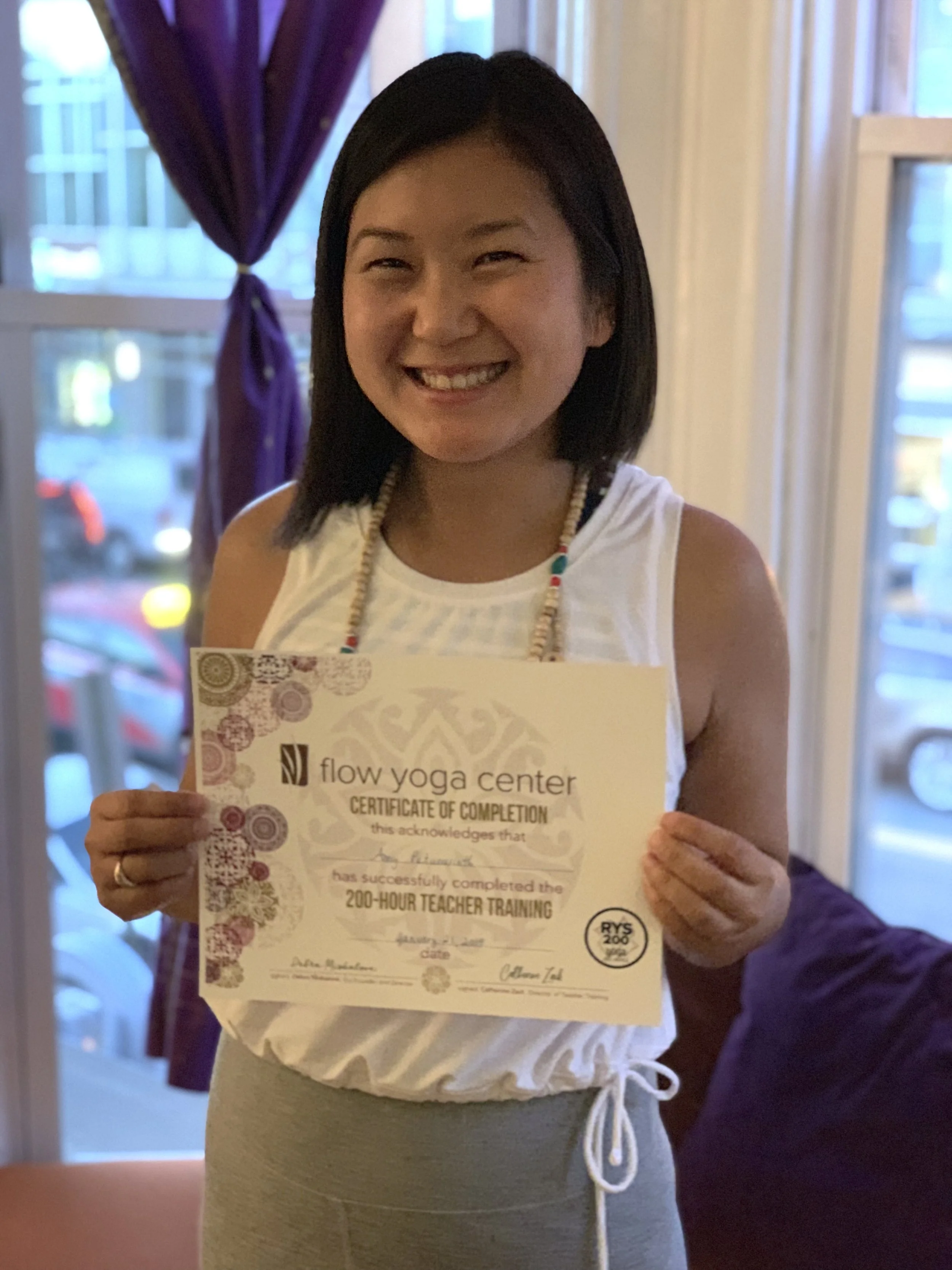 A woman with shoulder-length black hair smiling and holding a certificate of completion from Flow Yoga Center, indicating she has completed a 200-hour teacher training. She is indoors, near a window with purple curtains.