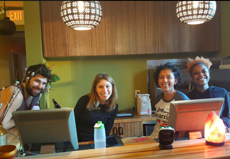 Four women sitting at a bar or counter, smiling and posing for the photo, with computer monitors and a salt lamp in front of them. Two hanging pendant lights are above them, and there is wooden cabinetry behind them.
