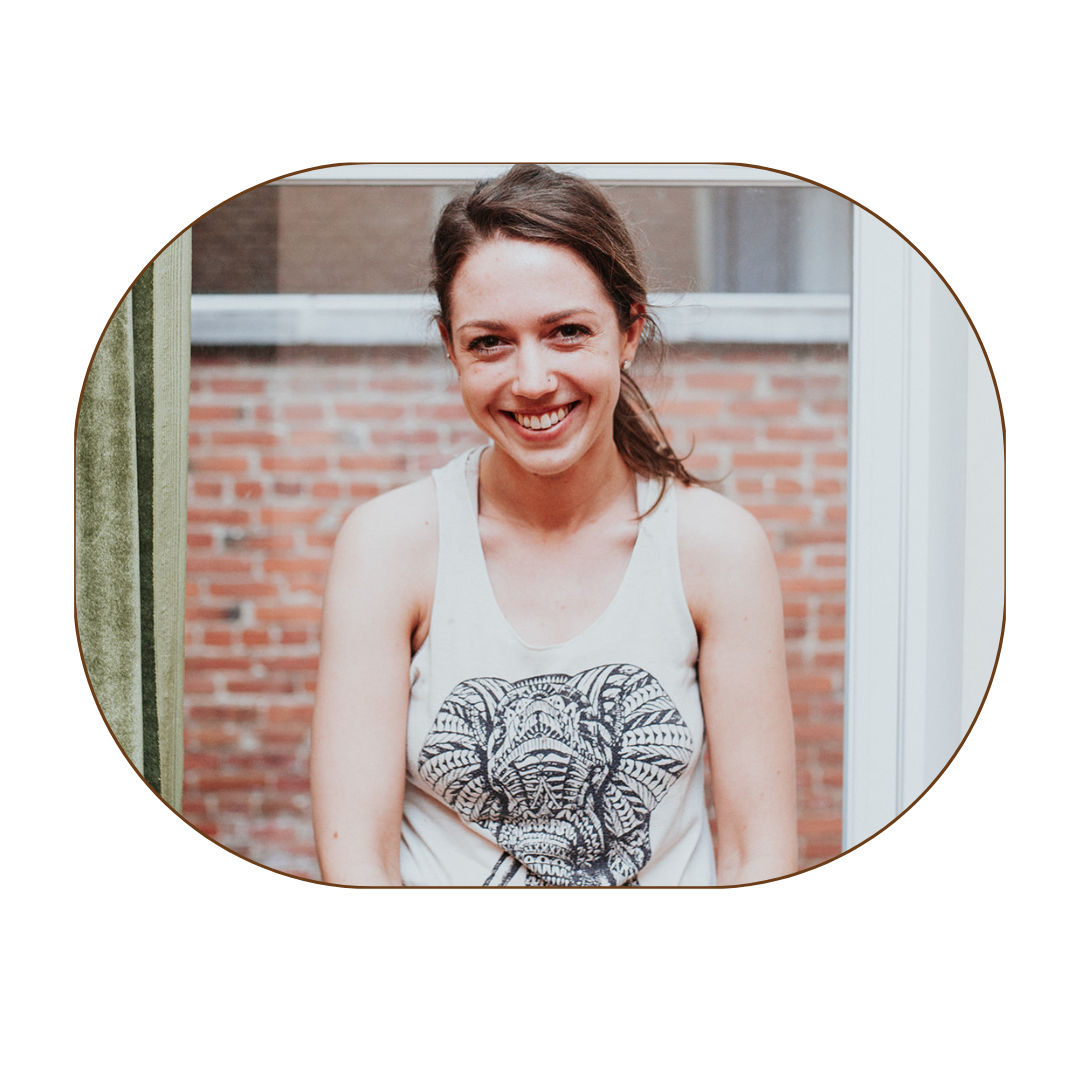 A young woman with brown hair, smiling, wearing a sleeveless top with a black and white animal print graphic, standing outdoors with a brick wall background.