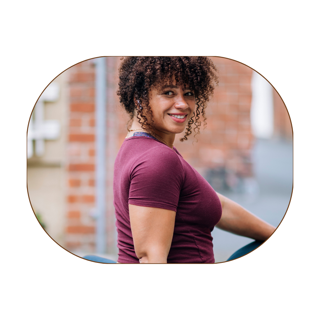 A woman with curly hair smiling, wearing a short-sleeved maroon shirt, standing outdoors with a brick wall and urban background.