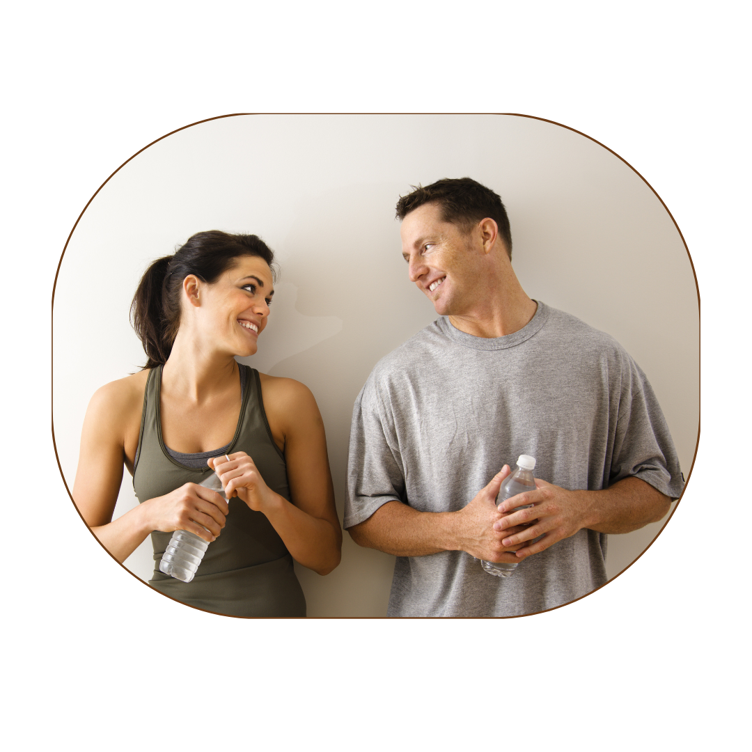 A man and woman smiling at each other, holding bottled water, and standing against a plain white wall.