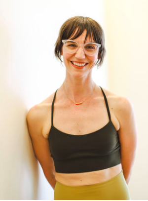 Woman with short brown hair, wearing pink glasses and a black spaghetti strap crop top, smiling in front of a plain light background.