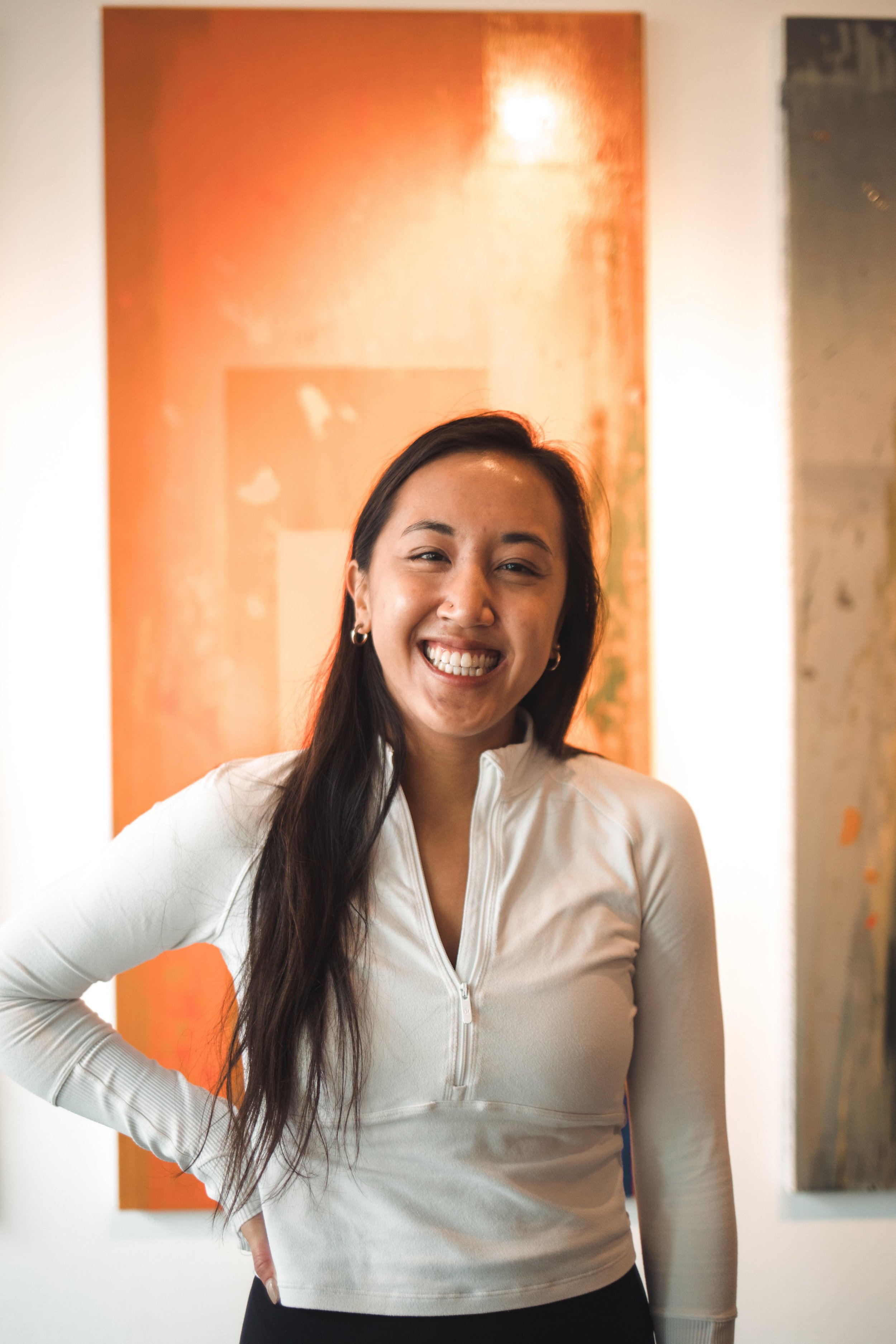 A smiling woman with long dark hair, wearing a white zip-up top, standing in front of colorful abstract paintings.
