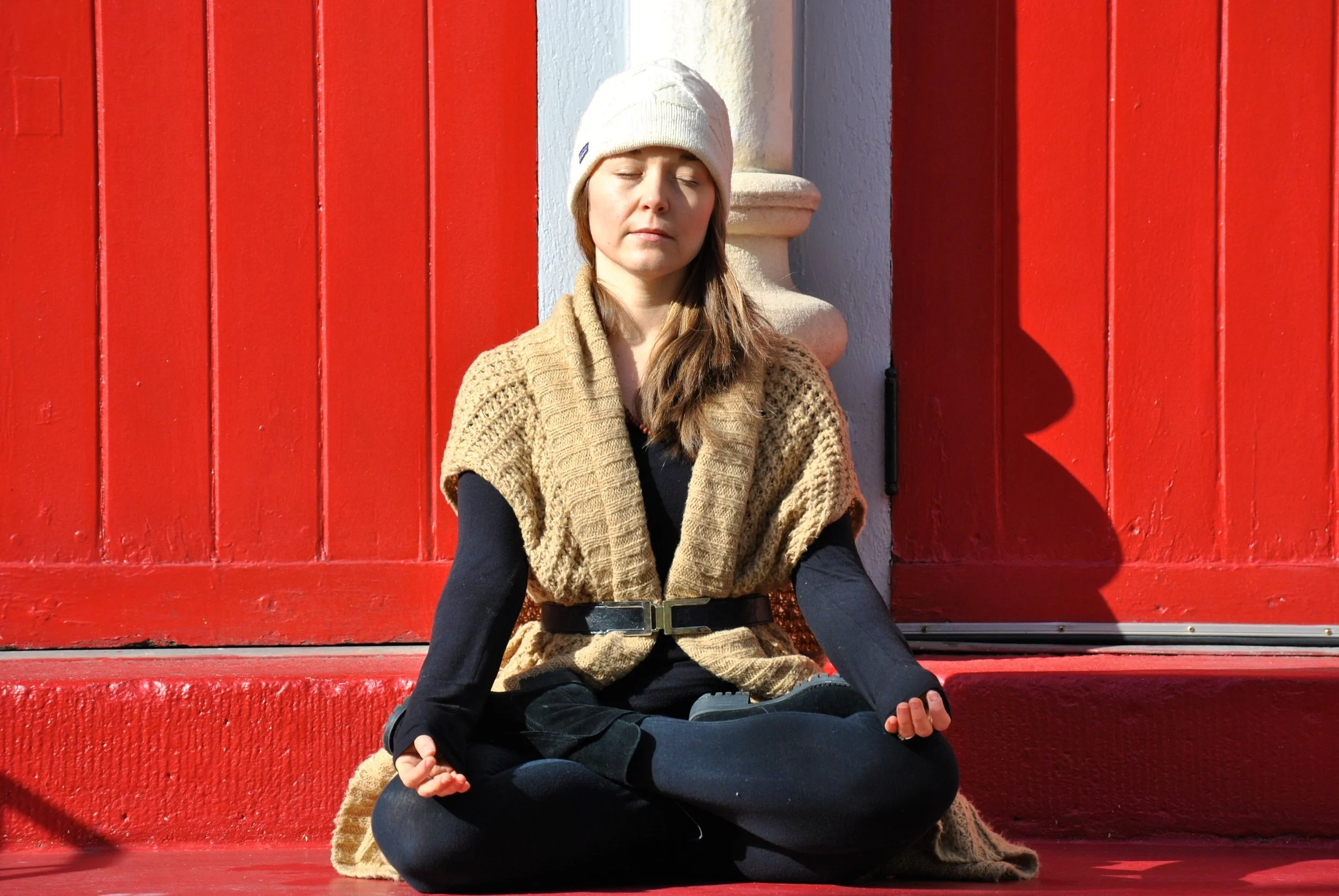 A young woman in a beige knit scarf, black long sleeves, and a white beanie is sitting cross-legged on red steps in a meditation pose with her eyes closed, near a red door.