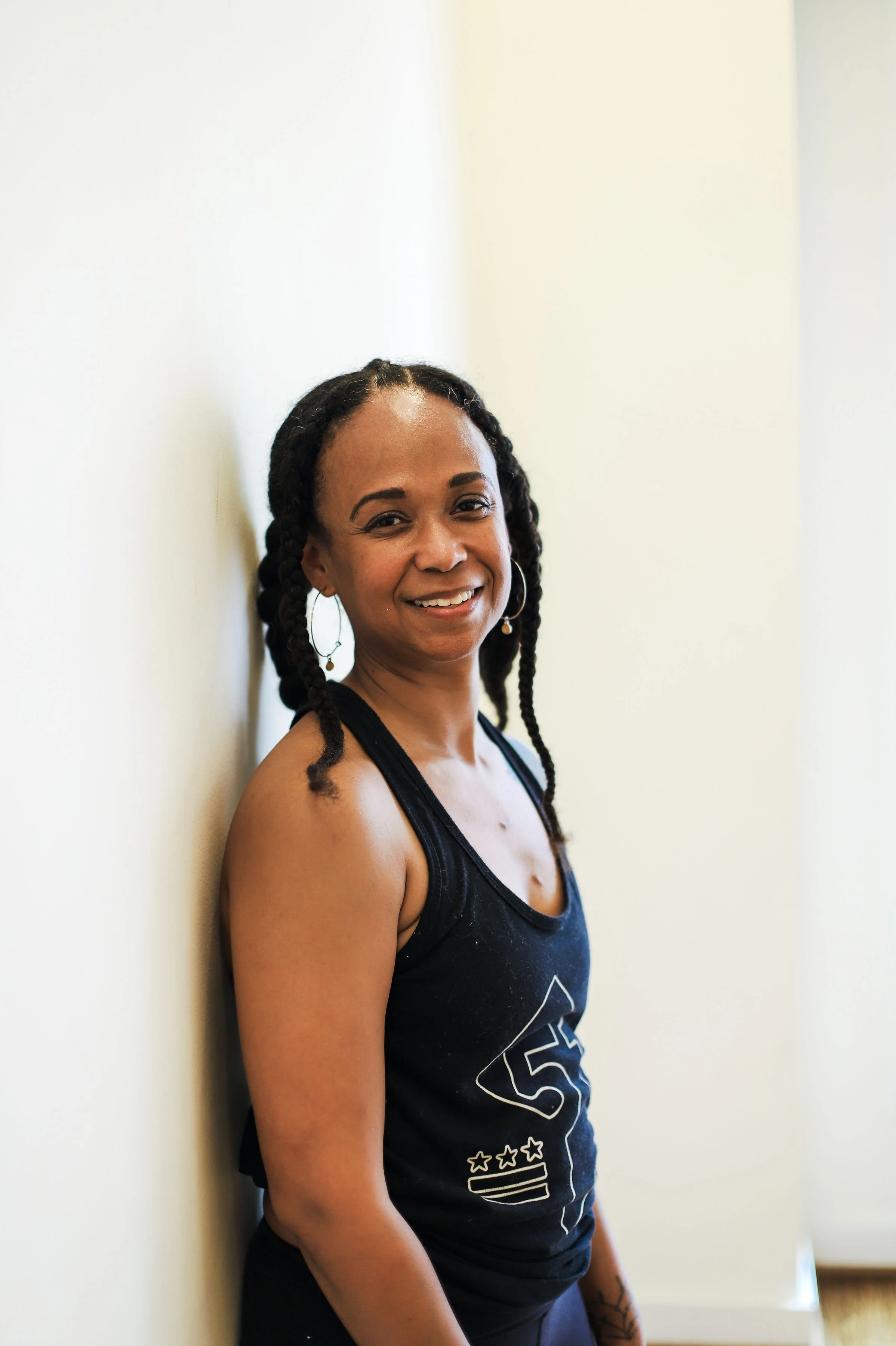 A woman who is a yoga teacher at Flow Yoga Center wearing hoop earrings and a black tank top, smiling while leaning against a light-colored wall.  