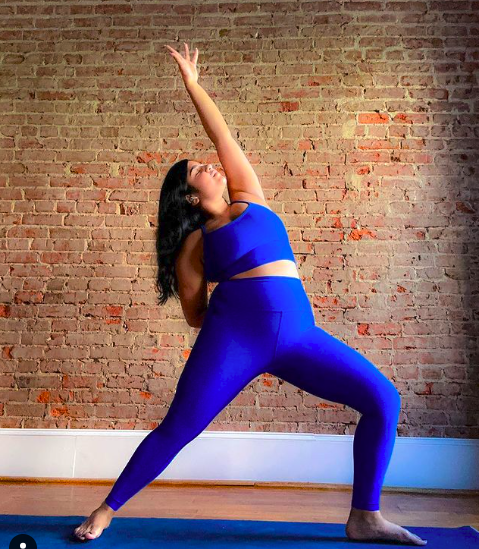 A woman practicing yoga in a blue athletic outfit against a brick wall, stretching with one arm raised and one leg bent.