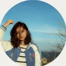 Young woman with shoulder-length hair posing outdoors against a blue sky, wearing a striped shirt and denim jacket with embroidered patches.