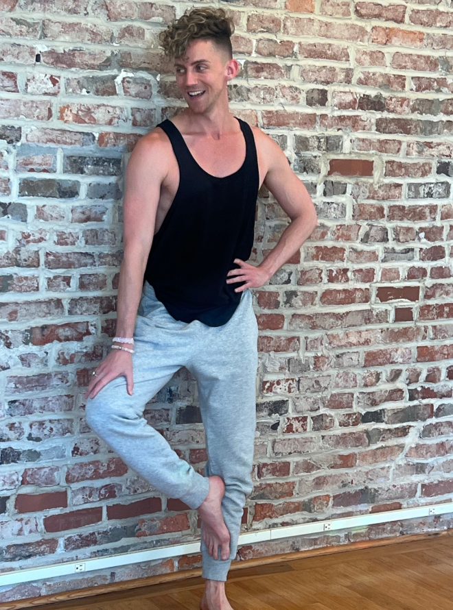 A person with curly hair wearing a black tank top and gray sweatpants stands against a brick wall, smiling and posing with one hand on their hip and the other resting on their thigh, barefoot at Flow Yoga Center. This person is a teacher.