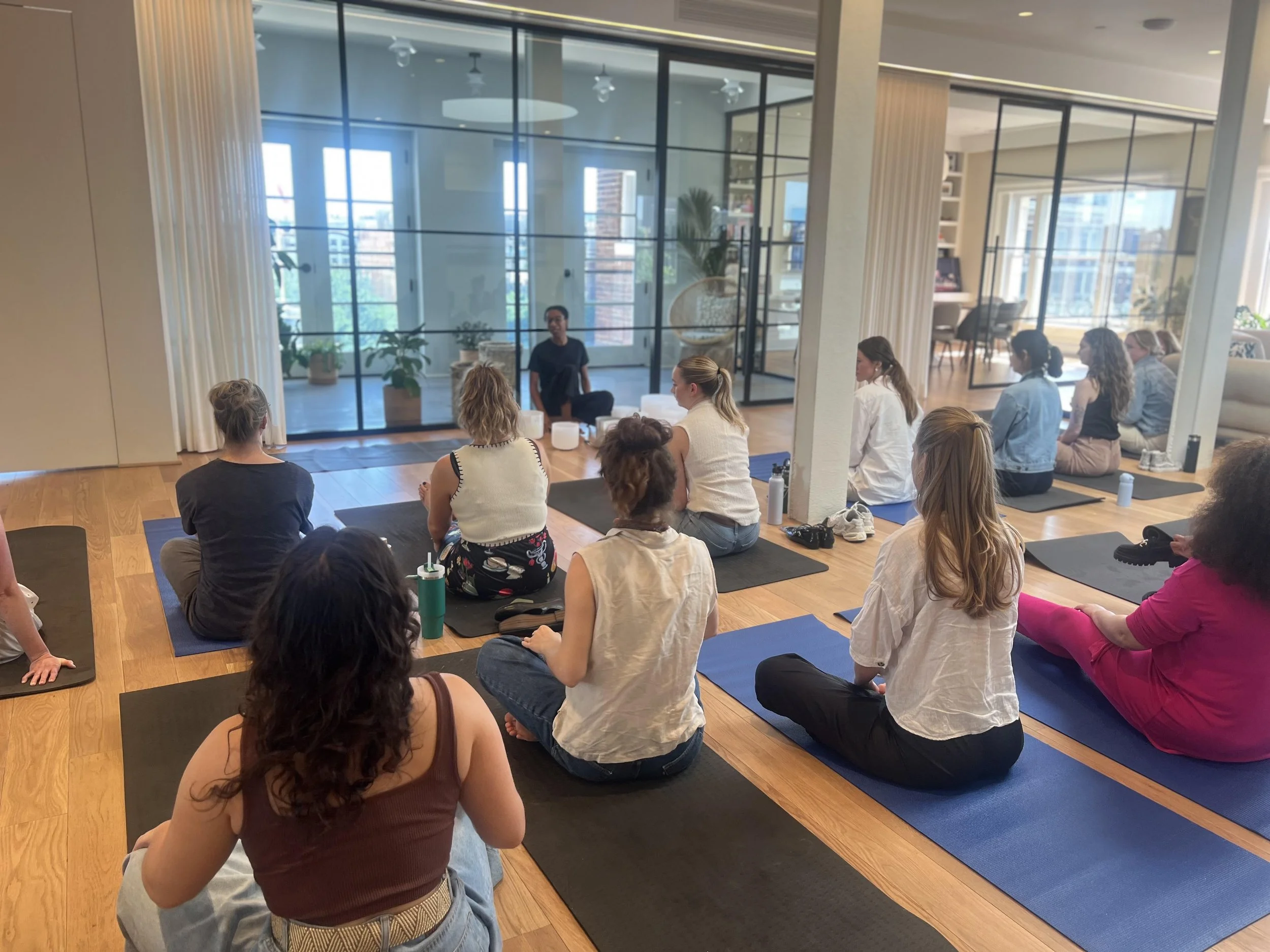 A group of people sitting on yoga mats in a yoga class or meditation session inside a modern, brightly lit room with large glass windows and a view of the city.