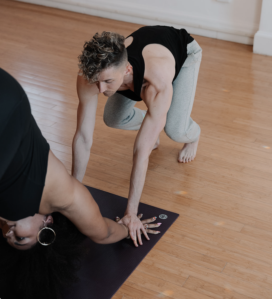 A man and woman practicing yoga on a wooden floor, with the man in a crouched position on his hands and knees, and the woman lying face down on her yoga mat with her eyes closed and mouth open.