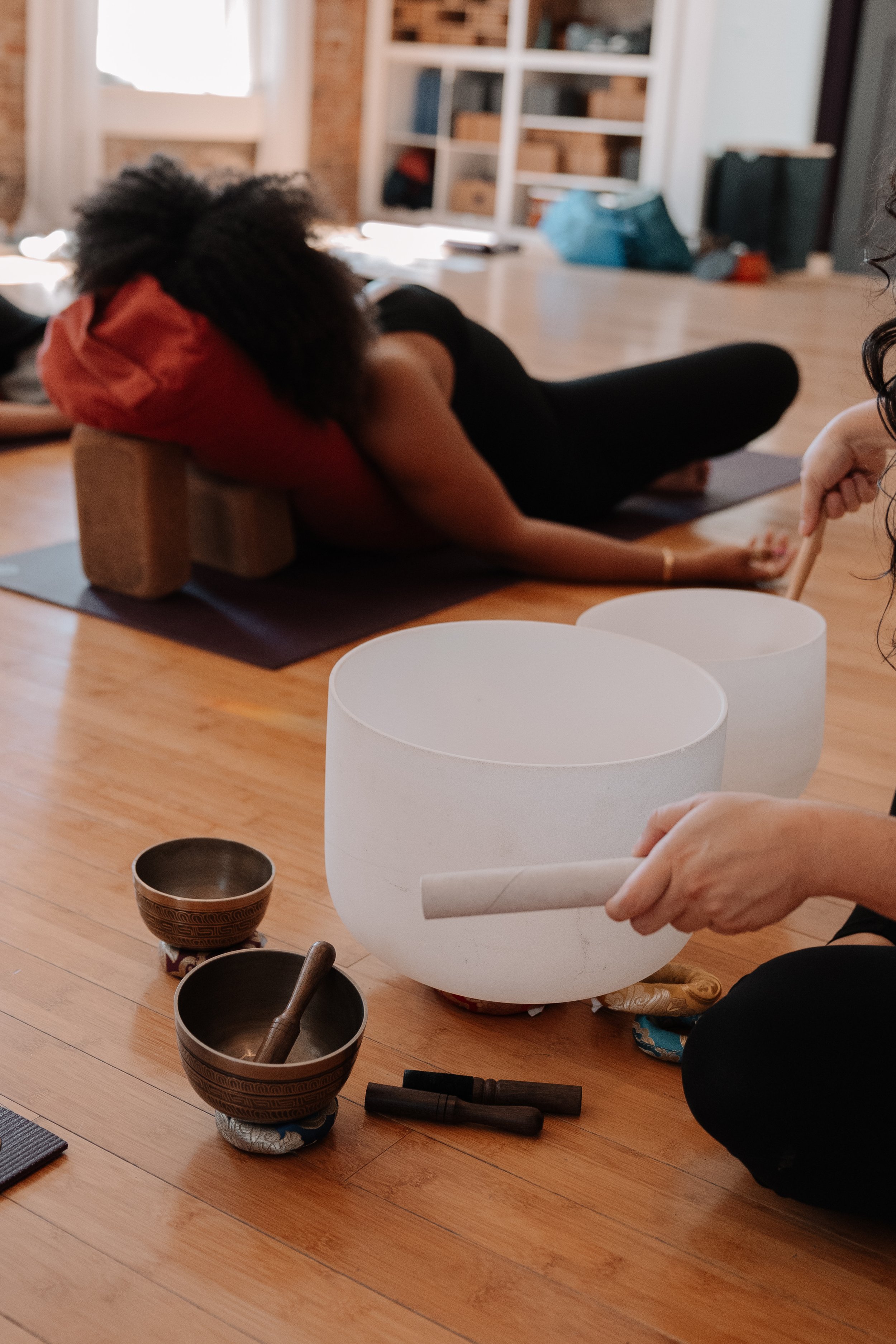 Person lying on yoga mat with head on cushion during a yoga session, with singing bowls on the floor in front.