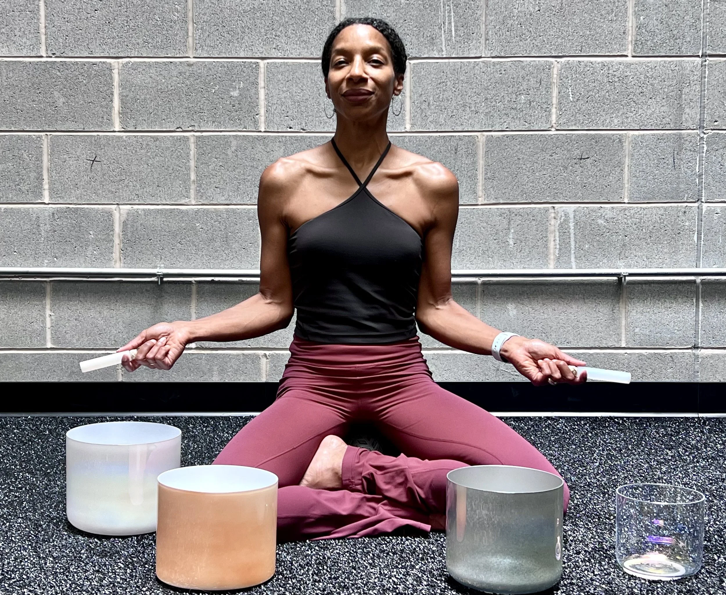 A woman in a black halter top and maroon pants practicing meditation, sitting cross-legged on a black textured surface in front of a gray brick wall, with singing bowls in front of her.