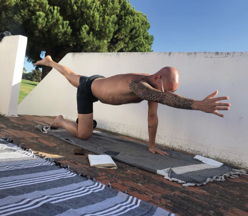 A man practicing yoga outdoors on a rooftop balcony, performing a bird dog pose on a gray yoga mat, with an open book nearby, trees in the background, and a white wall behind him.