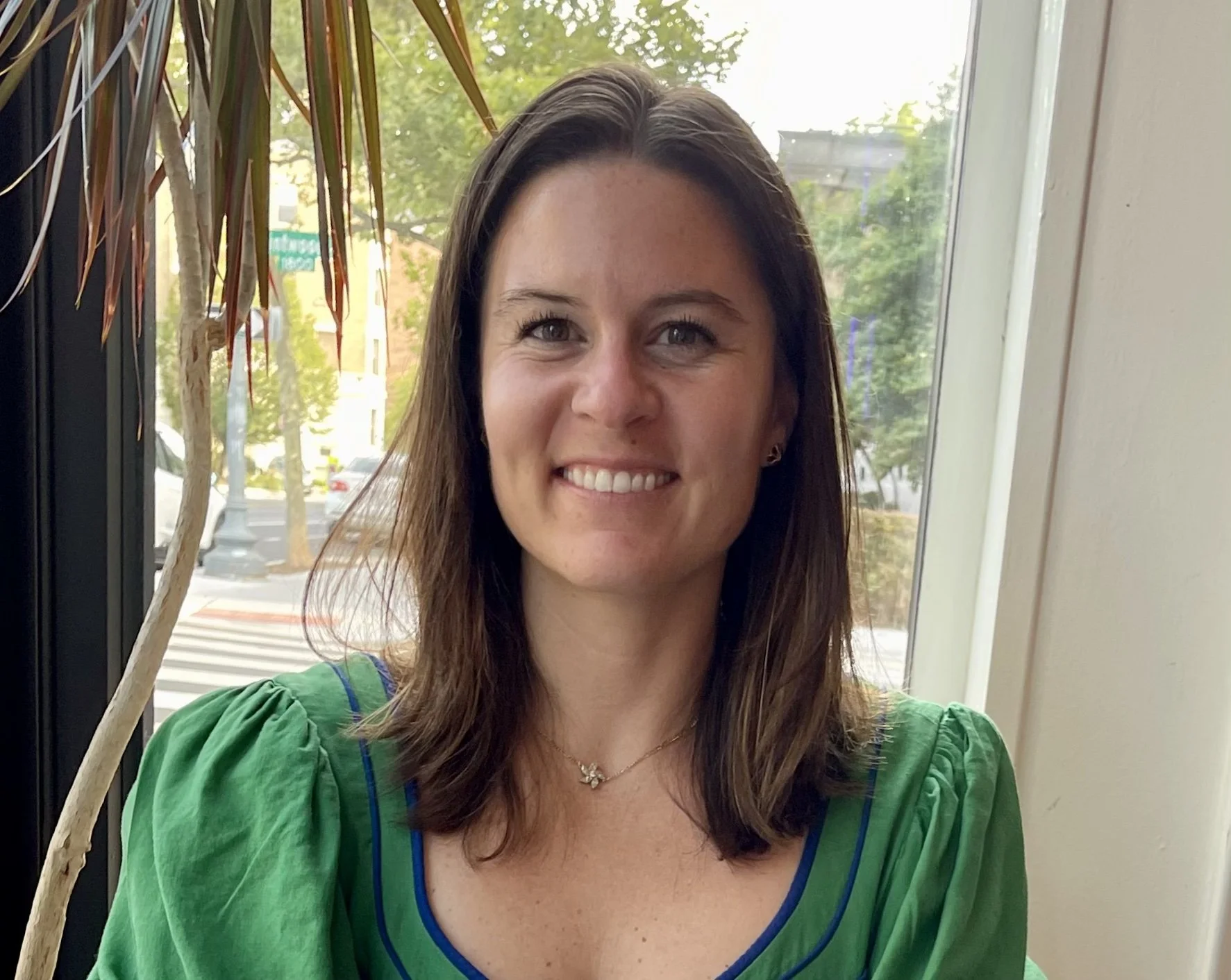 A woman with shoulder-length brown hair, wearing a green top with puffed sleeves, sitting near a window with a view of trees and street outside.