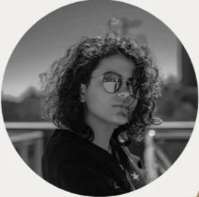 Black and white photo of a woman with curly hair wearing sunglasses, outdoors on a sunny day.