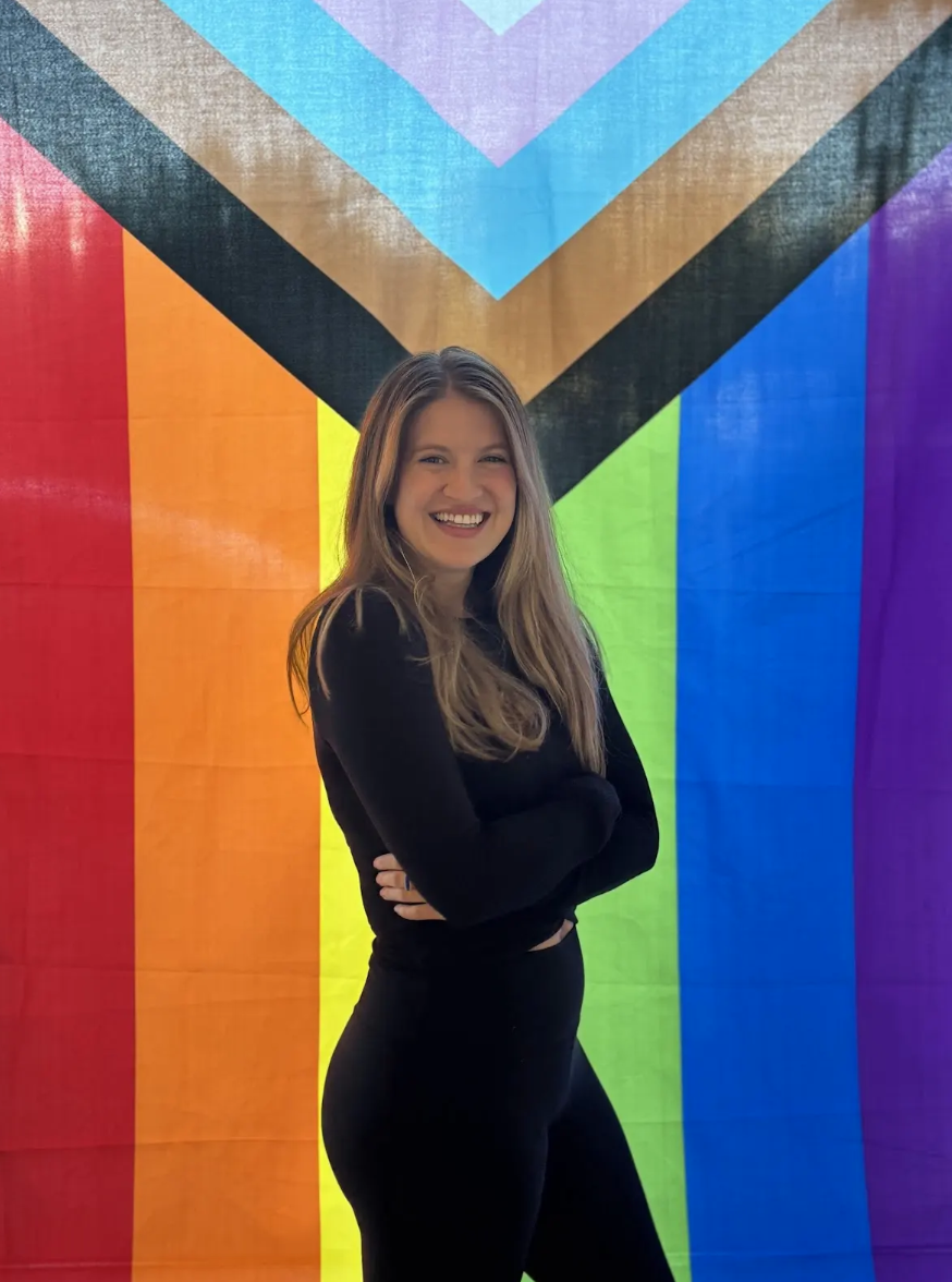A young woman with long blonde hair is smiling with her arms crossed in front of a colorful pride flag with rainbow stripes and a black, brown, light blue, pink, and white chevron.