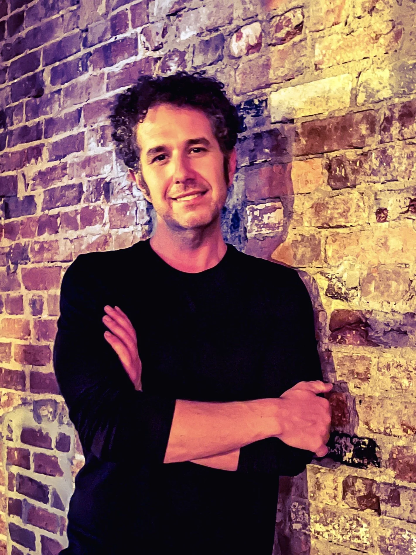 A man with curly hair, wearing a black long sleeve shirt, standing with arms crossed in front of a brick wall.