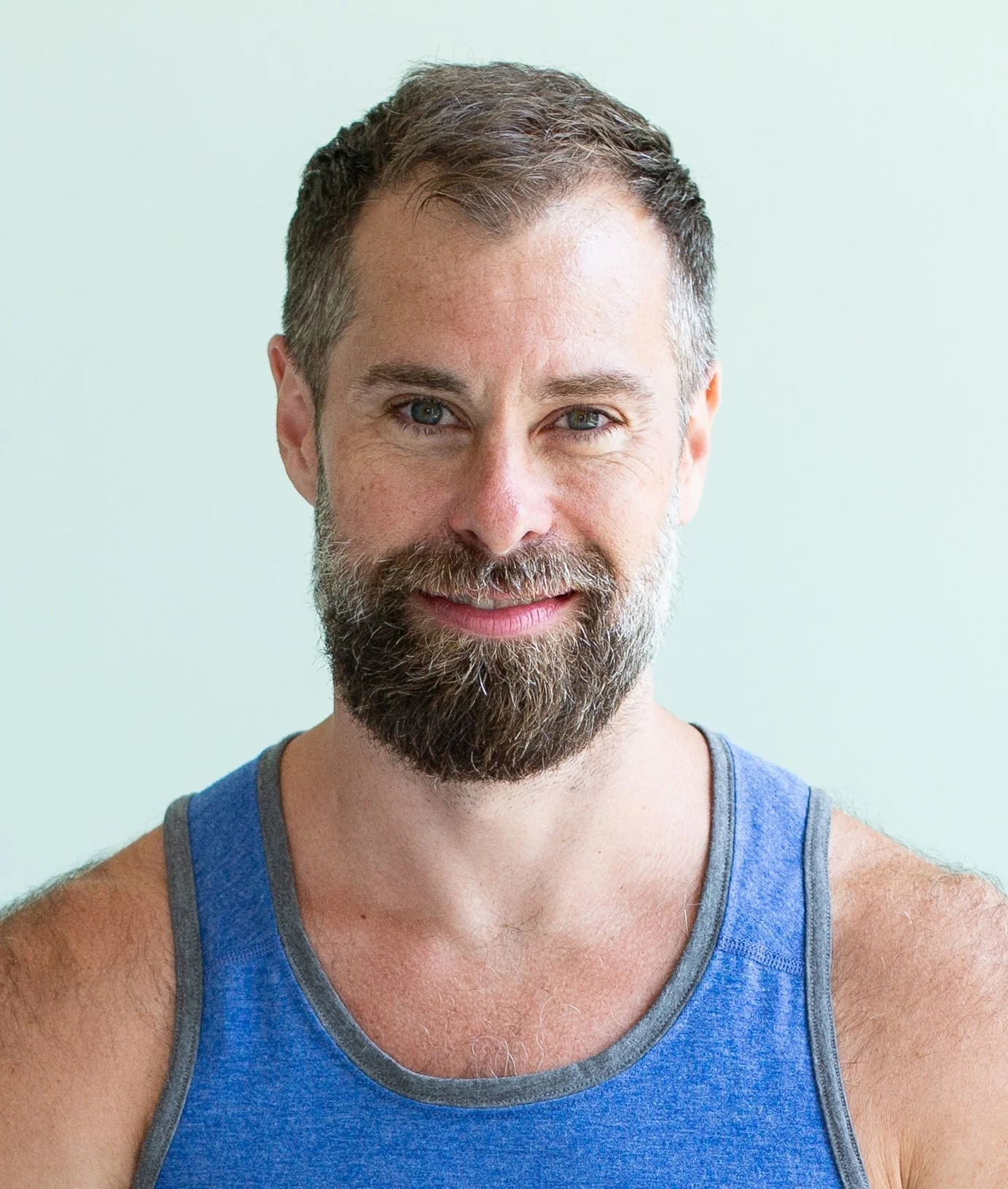 A smiling man with a beard and short hair wearing a blue tank top, standing against a light green background.