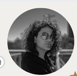 Black and white photo of a young woman with curly hair, wearing sunglasses, standing outdoors with a blurred cityscape in the background.