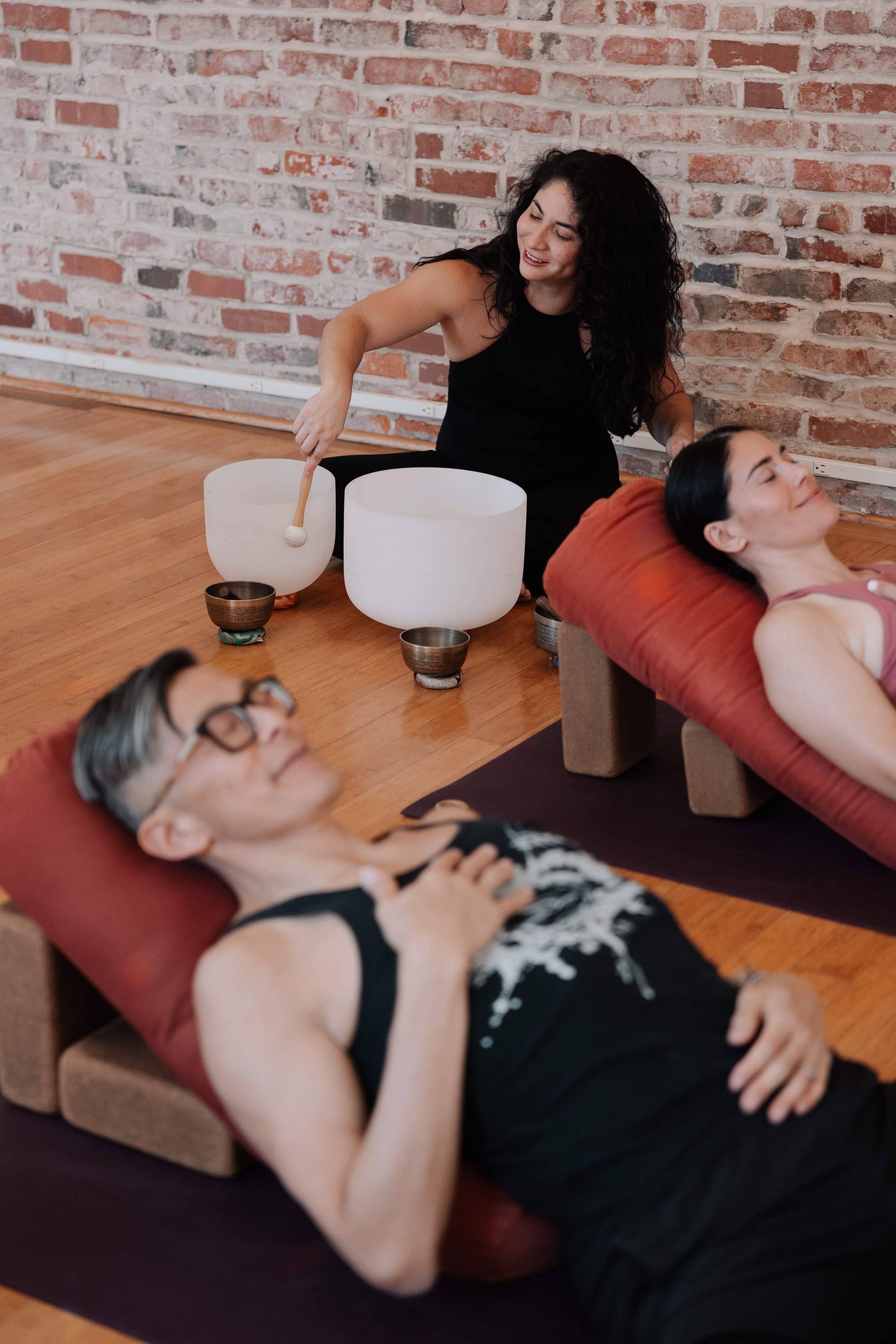 A woman is playing crystal singing bowls during a sound healing session with two people lying on massage tables with cushions, in a room with a brick wall.