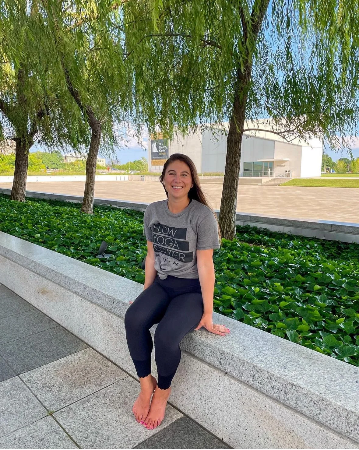 A woman with long brown hair smiling and sitting barefoot on a stone ledge outdoors, with lush green trees and a modern white building in the background.