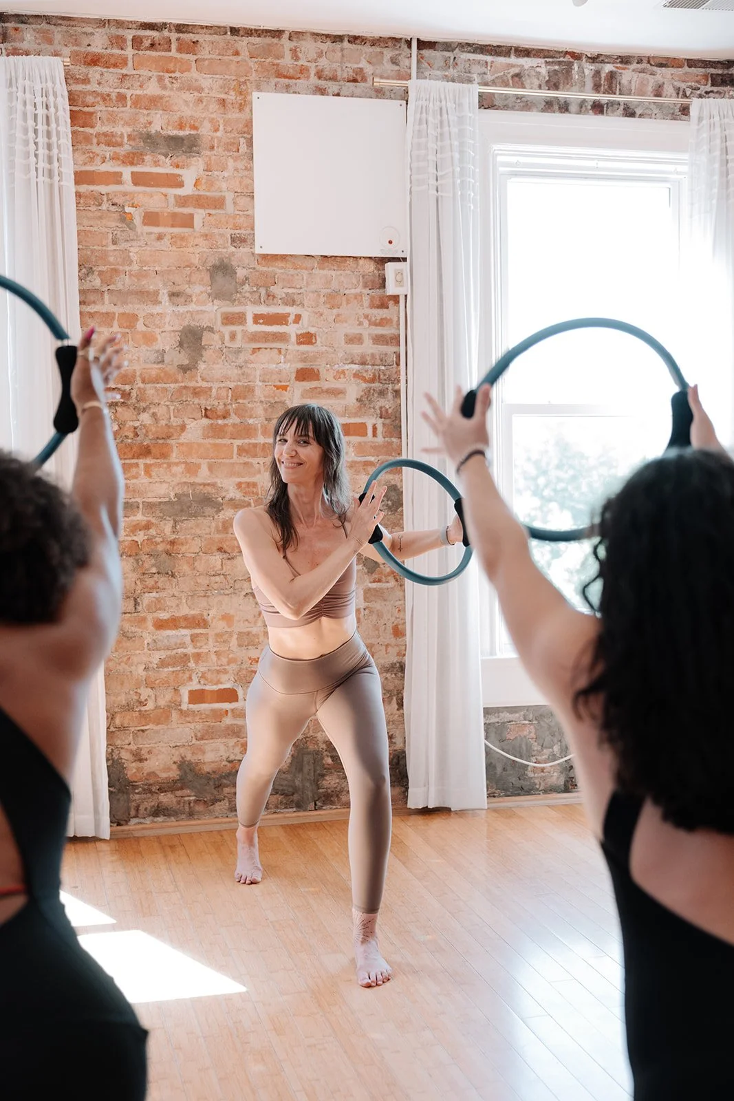 A woman leading a fitness or dance class using hoops, with two participants holding hoops in a brightly lit studio with a brick wall and large windows.