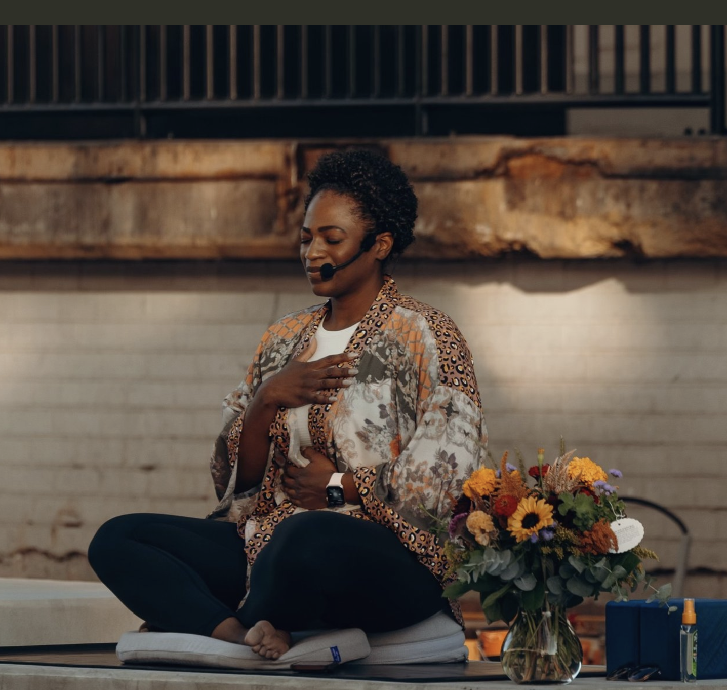 A woman meditating on a cushion with her hand on her chest and her eyes closed, sitting cross-legged on a platform with a flower arrangement nearby, in what appears to be a yoga or meditation space.