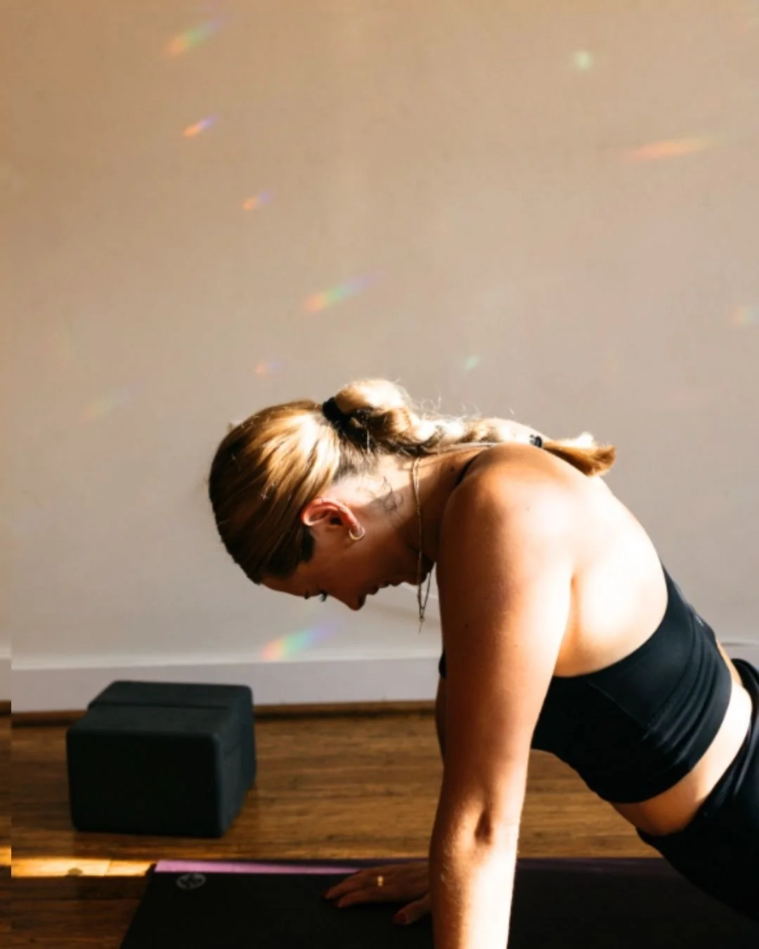 A woman practicing yoga indoors on a black mat, with rainbow light reflections on the wall behind her.