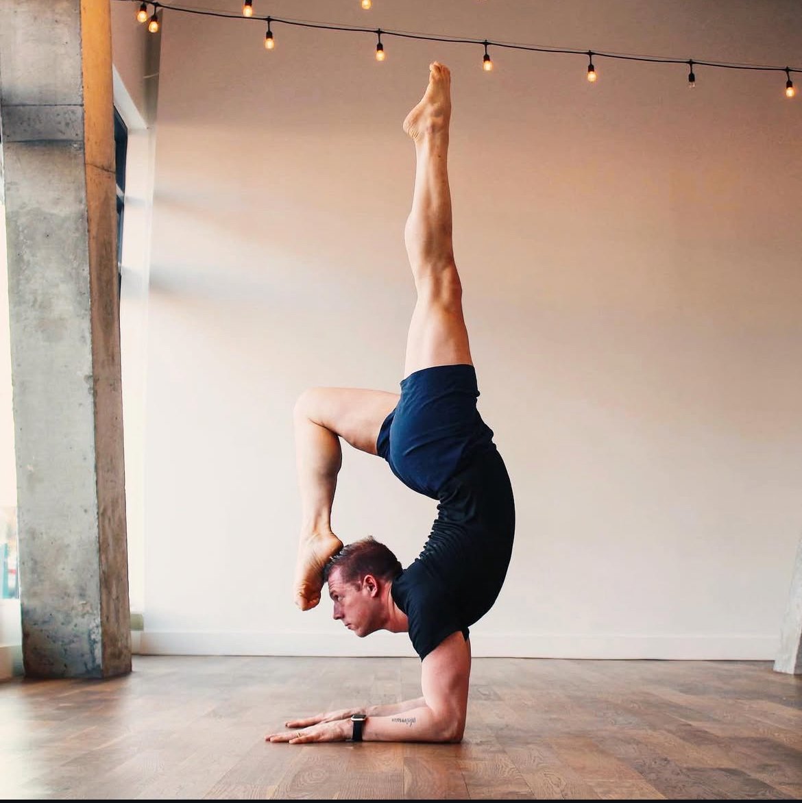 Man performing a headstand yoga pose with one leg extended upwards and the other bent, on a wooden floor in a room with string lights.
