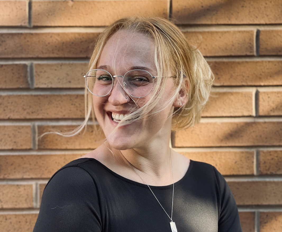 Smiling woman with blonde hair, glasses, and a necklace standing in front of a brick wall.