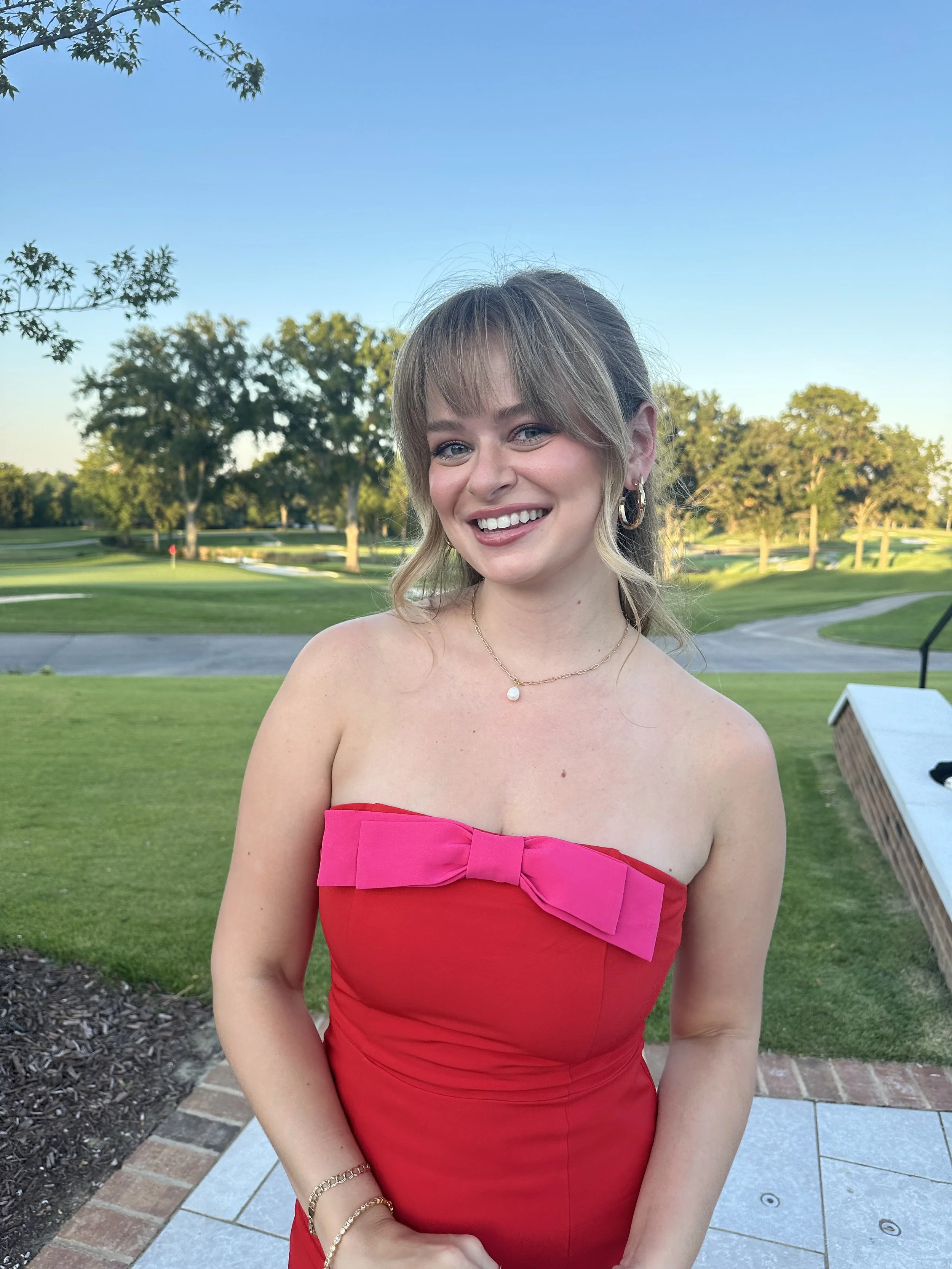A smiling young woman in a strapless red dress with a pink bow, standing outdoors on a sunny day with a golf course and trees in the background.