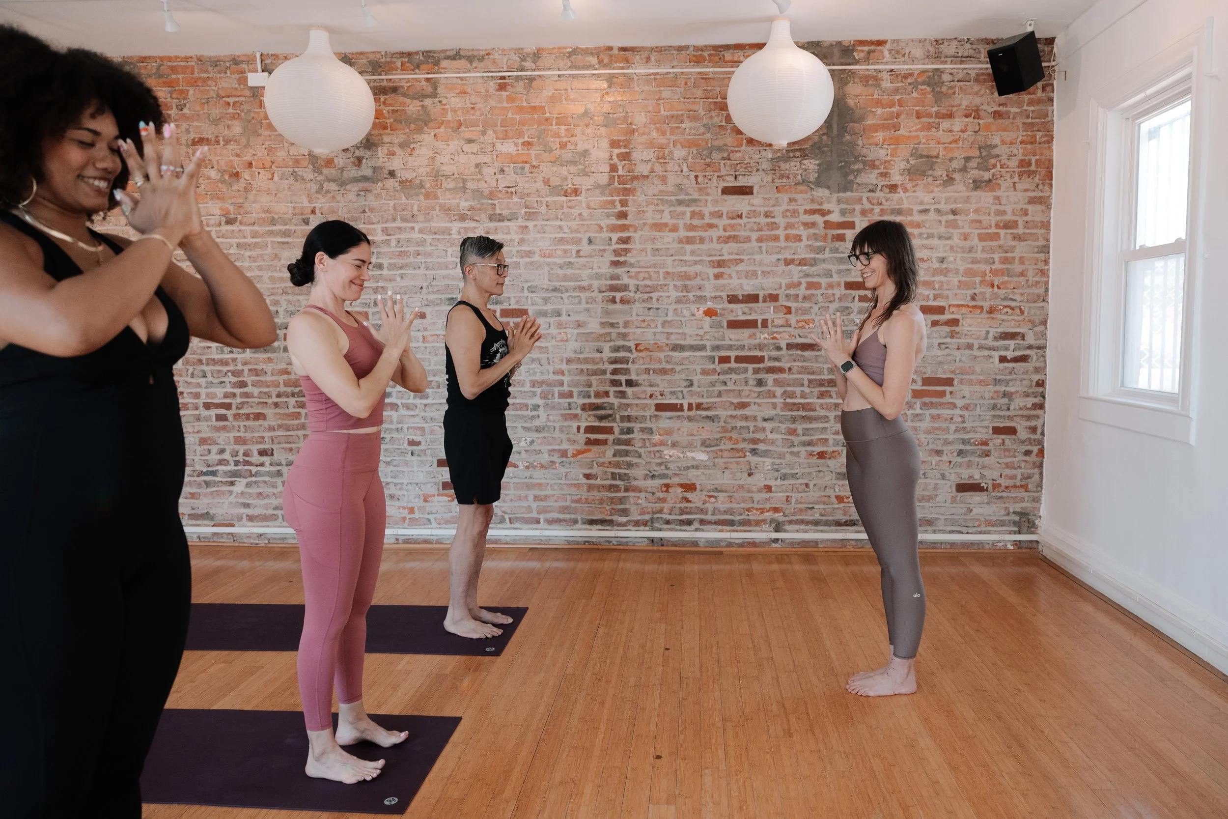 Five women participating in a yoga or meditation class in a room with a brick wall, hardwood floor, and white paper lantern lights. Four women are standing on yoga mats with hands in prayer position, facing a woman at the front who is leading the session.