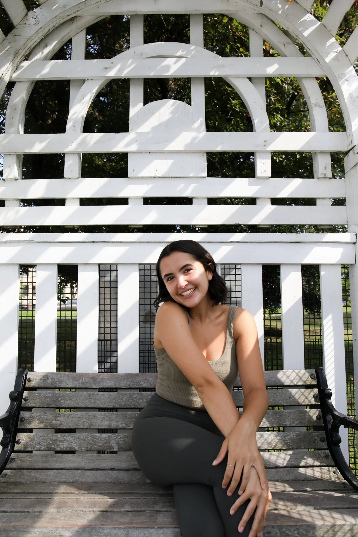 A young woman with short dark hair smiling while sitting on a wooden bench outdoors, with a white decorative lattice behind her and greenery visible through the lattice.