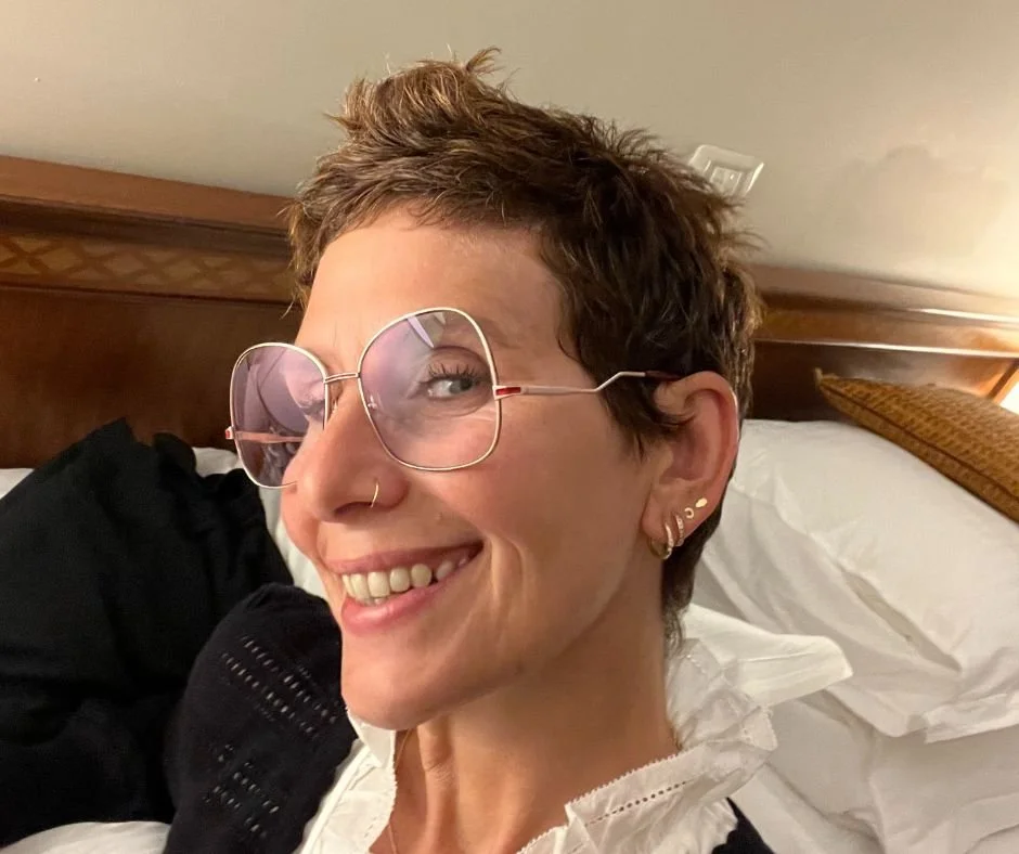 Smiling woman with short, wavy brown hair, wearing large, round pink-tinted glasses and multiple earrings, lying on a bed in a wooden-paneled room.