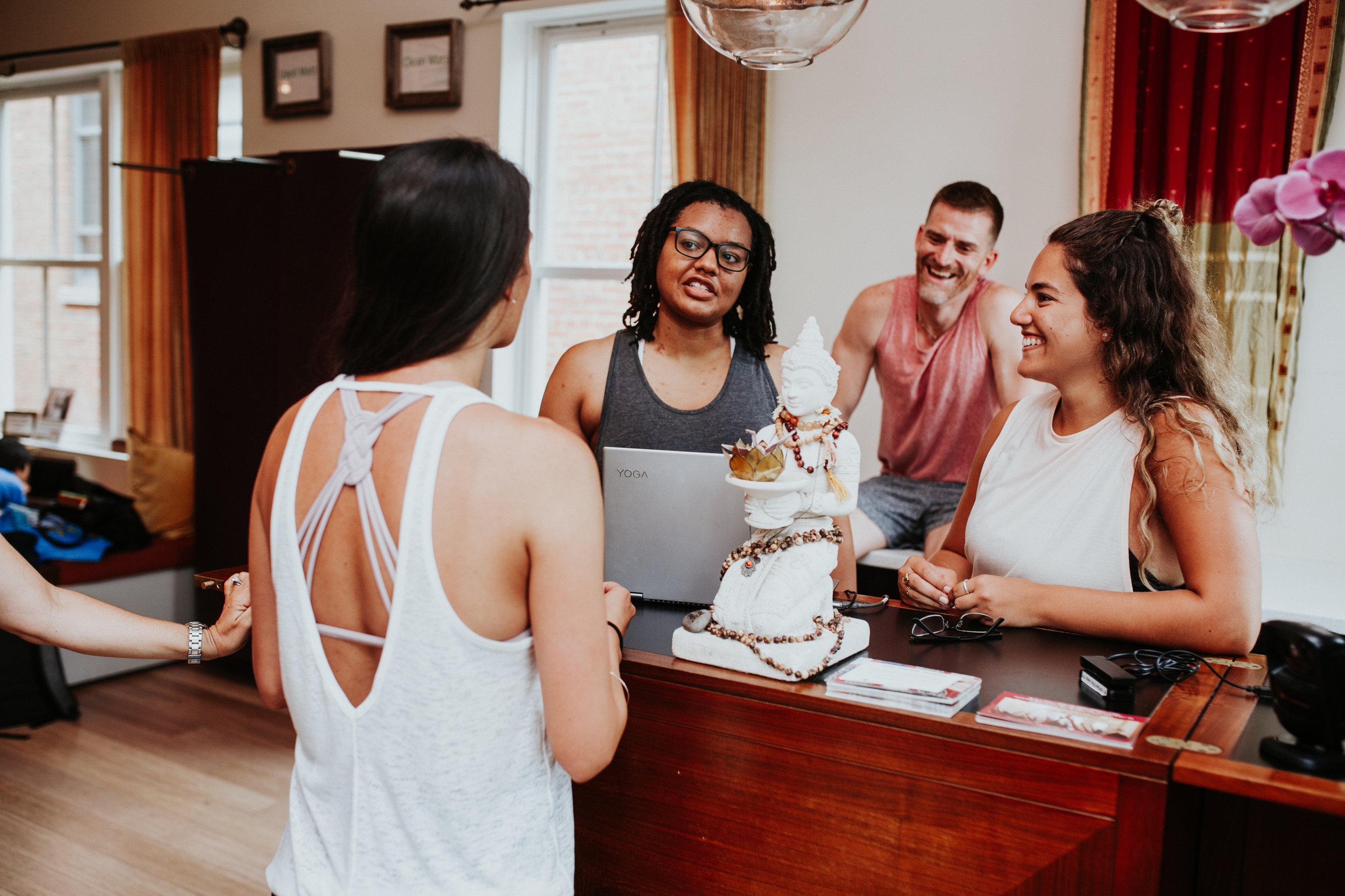 Four people engaged in a conversation around a wooden reception desk in a well-lit indoor space. One woman has an interesting backless top with crisscross straps, and another woman is wearing glasses and a sleeveless top. A smiling man and woman are on the other side of the desk. The desk has religious or decorative items, such as a small statue and rosary beads, along with some brochures and electronic cords.