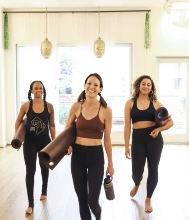 Three women in athletic clothing smiling and walking with yoga mats inside a bright room during a fitness class.