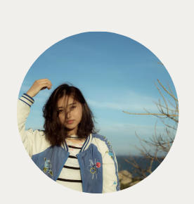 Young woman with shoulder-length dark hair, wearing a striped shirt and a blue jacket with floral embroidery, standing outdoors against a clear blue sky.