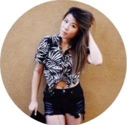 Young woman with long hair wearing a patterned button-up shirt and black shorts, standing against a tan background.