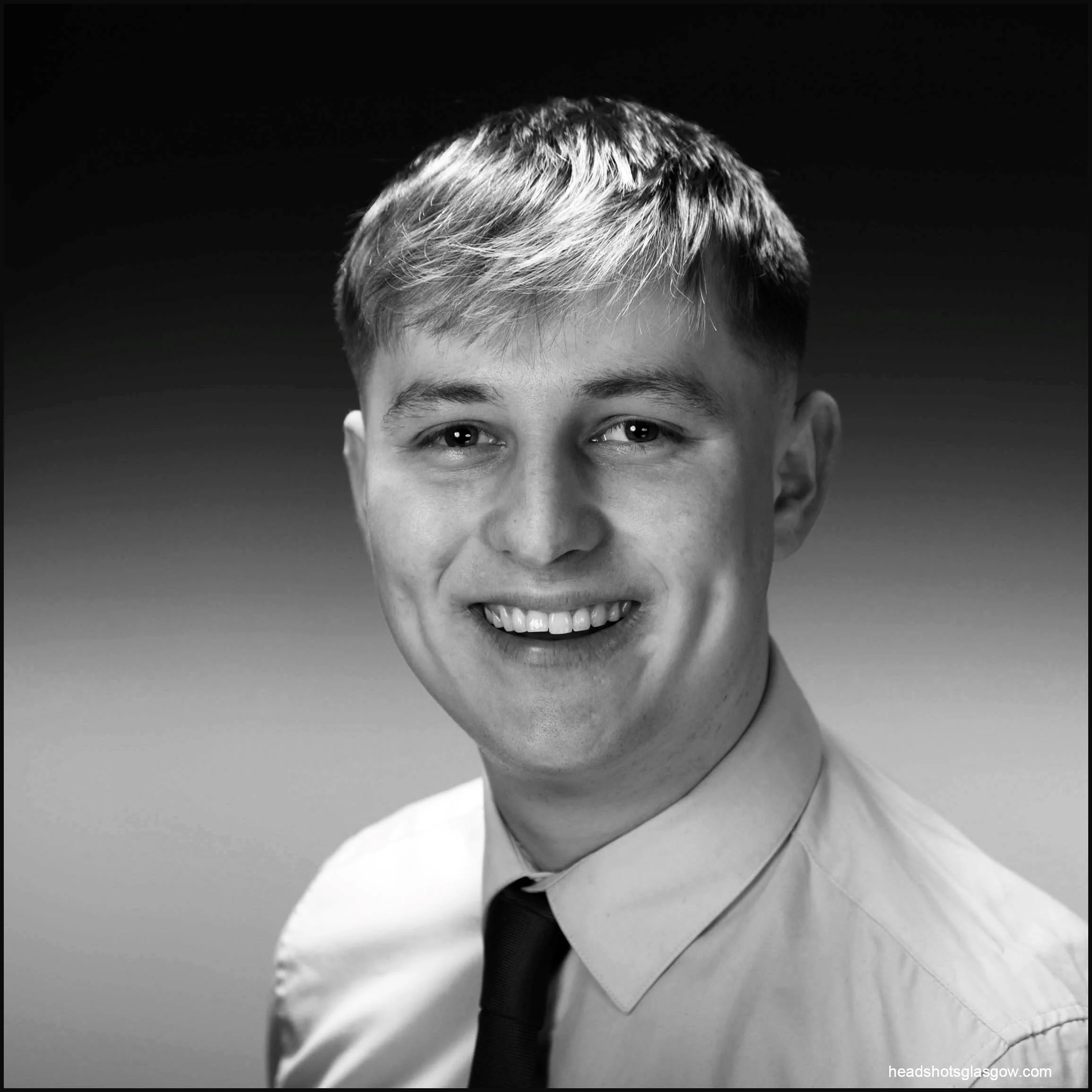 Black and white portrait of a young man smiling, wearing a collared shirt and tie, with a blurred gradient background.